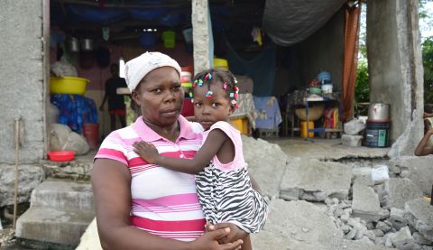 Tessa, a mother of 10, stands in front of her ruined house in Haiti holding her daughter in her arms Photo: Fabienne Douce/ActionAid.