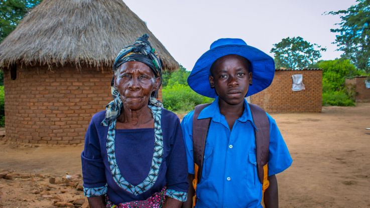 Mutsawashe and his grandmother stand outside a house in Zimbabwe.